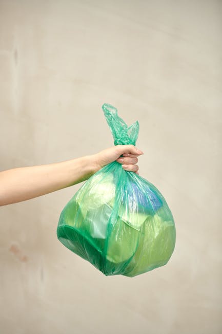 A person’s arm extended into the frame, holding a green and blue plastic garbage bag tightly by its twisted top. The bag is translucent, showing the contents inside, and appears full, with slightly bu