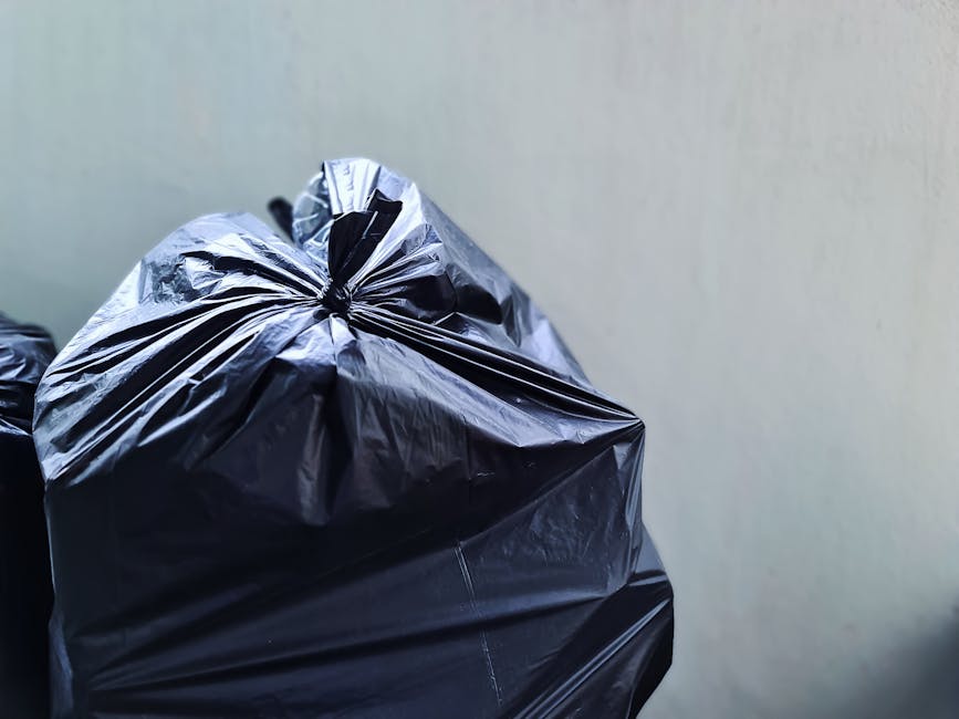 A person’s arm extended into the frame, holding a green and blue plastic garbage bag tightly by its twisted top. The bag is translucent, showing the contents inside, and appears full, with slightly bu