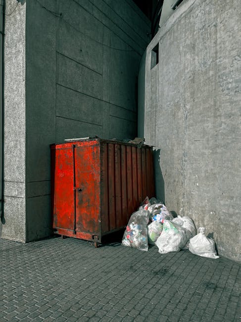 A large, upright metal waste container with a weathered, red-painted finish showing signs of rust and paint chipping, positioned against a dark, textured wall on the left side of the image. The contai